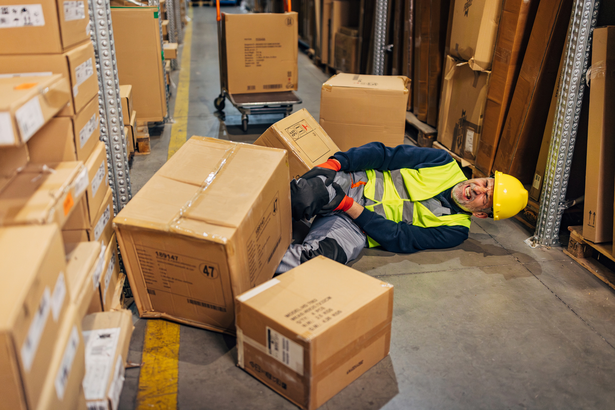 Heavy cardboard boxes fell from a shelf and knocked down a middle age warehouse worker in reflective uniform