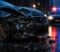 personal-injury-representation Damaged dark sedan with a crumpled front end on a wet city street at night, police lights flashing in the background.