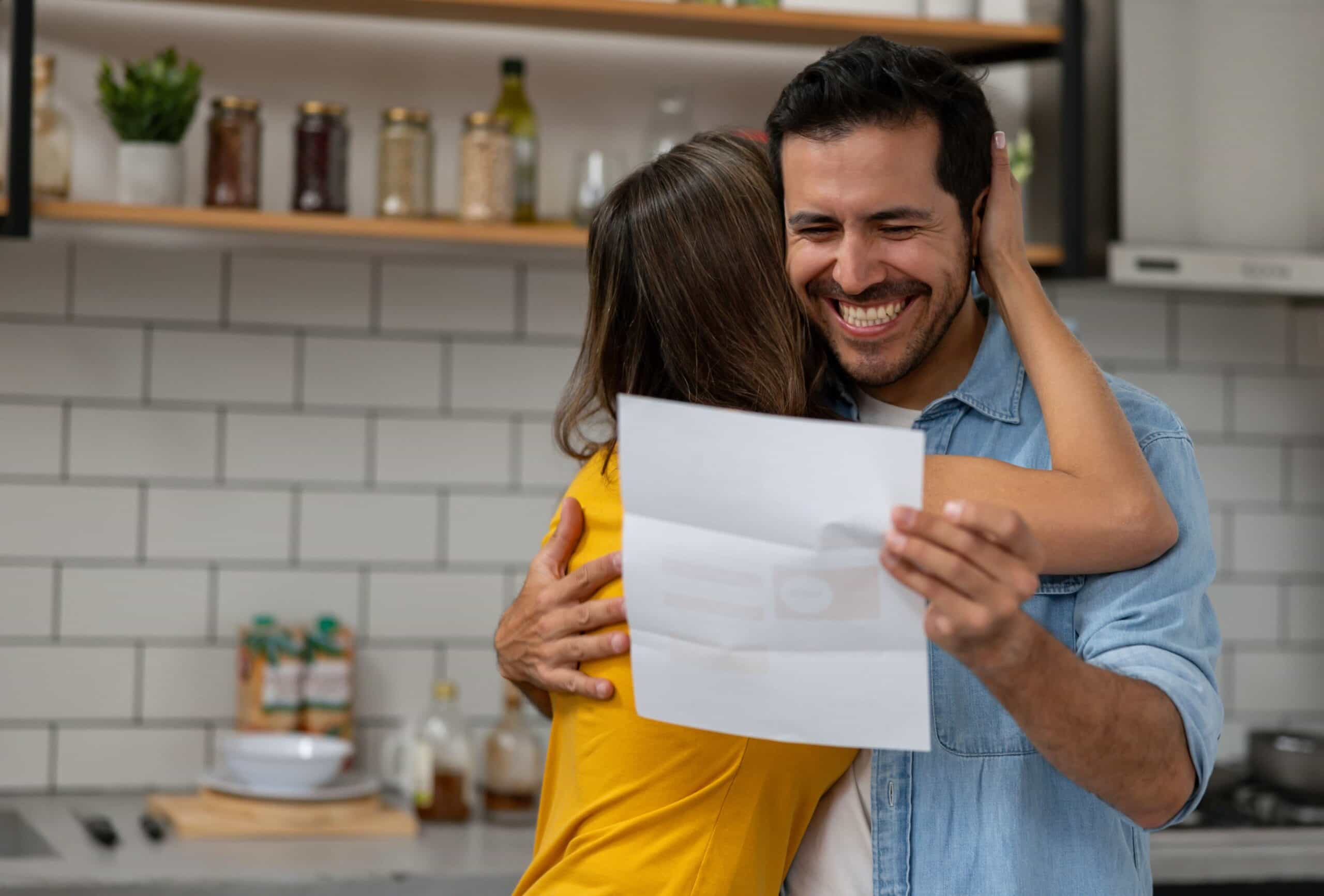 A joyful embrace between a man in a denim shirt and a woman in yellow, holding a letter together in a bright kitchen.